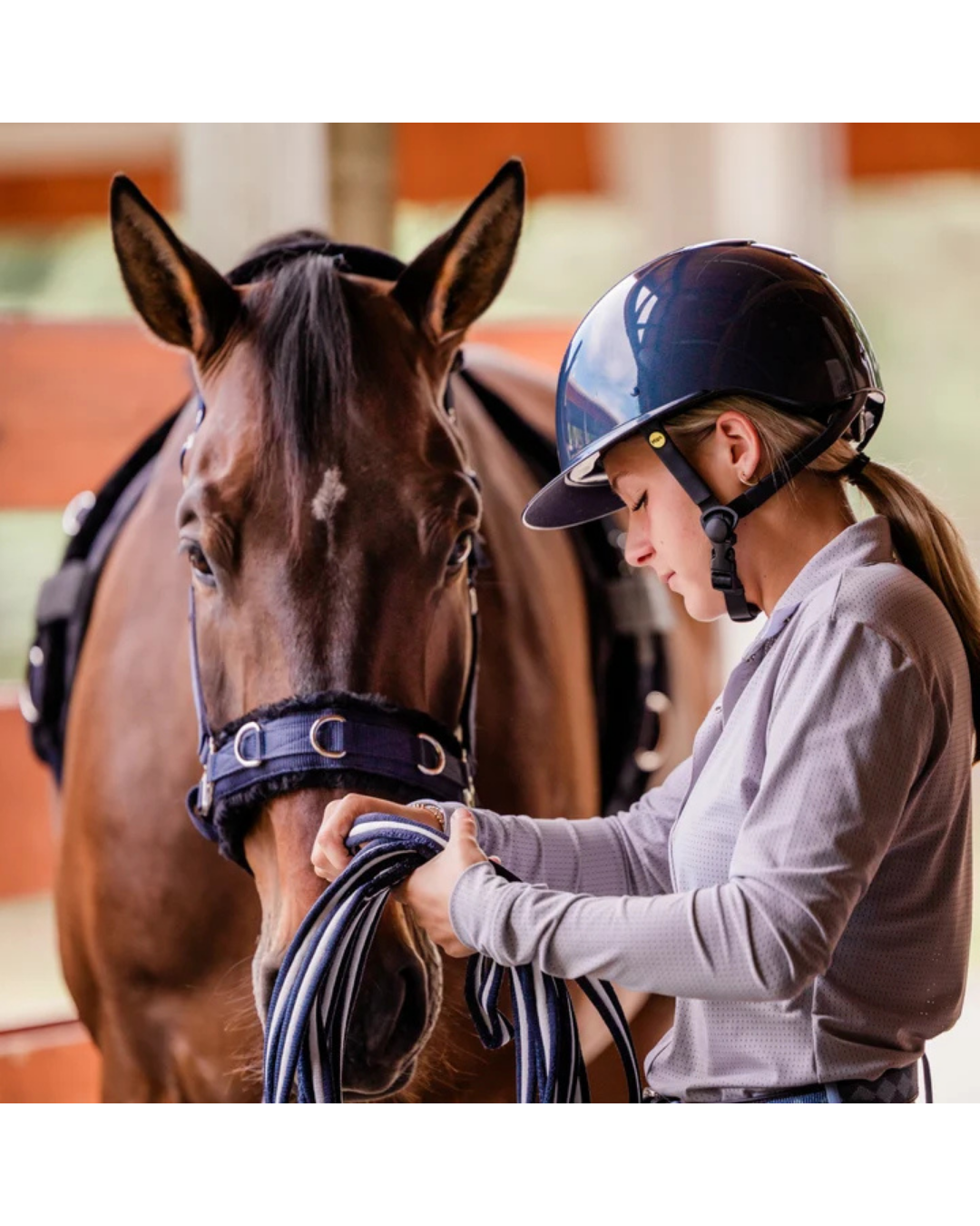 girl with navy blue shiny glossy helmet with black harness straps and yellow mips logo fixing a lunge line on a horse equestrian fashion outfitters