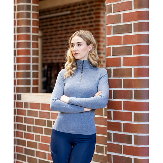 A woman stands outdoors against a red brick column, wearing a heathered blue mock-neck long-sleeve quarter-zip pullover and dark navy tights equestrian fahsion outfitters