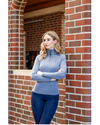 A woman stands outdoors against a red brick column, wearing a heathered blue mock-neck long-sleeve quarter-zip pullover and dark navy tights equestrian fahsion outfitters