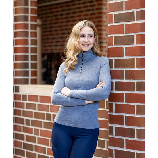 A woman with blonde hair stands leaning against a red brick wall, wearing a heathered blue mock neck  1/4 zip long-sleeve top and dark blue tights equestrian fashion outfitters