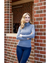A woman with blonde hair stands leaning against a red brick wall, wearing a heathered blue mock neck  1/4 zip long-sleeve top and dark blue tights equestrian fashion outfitters