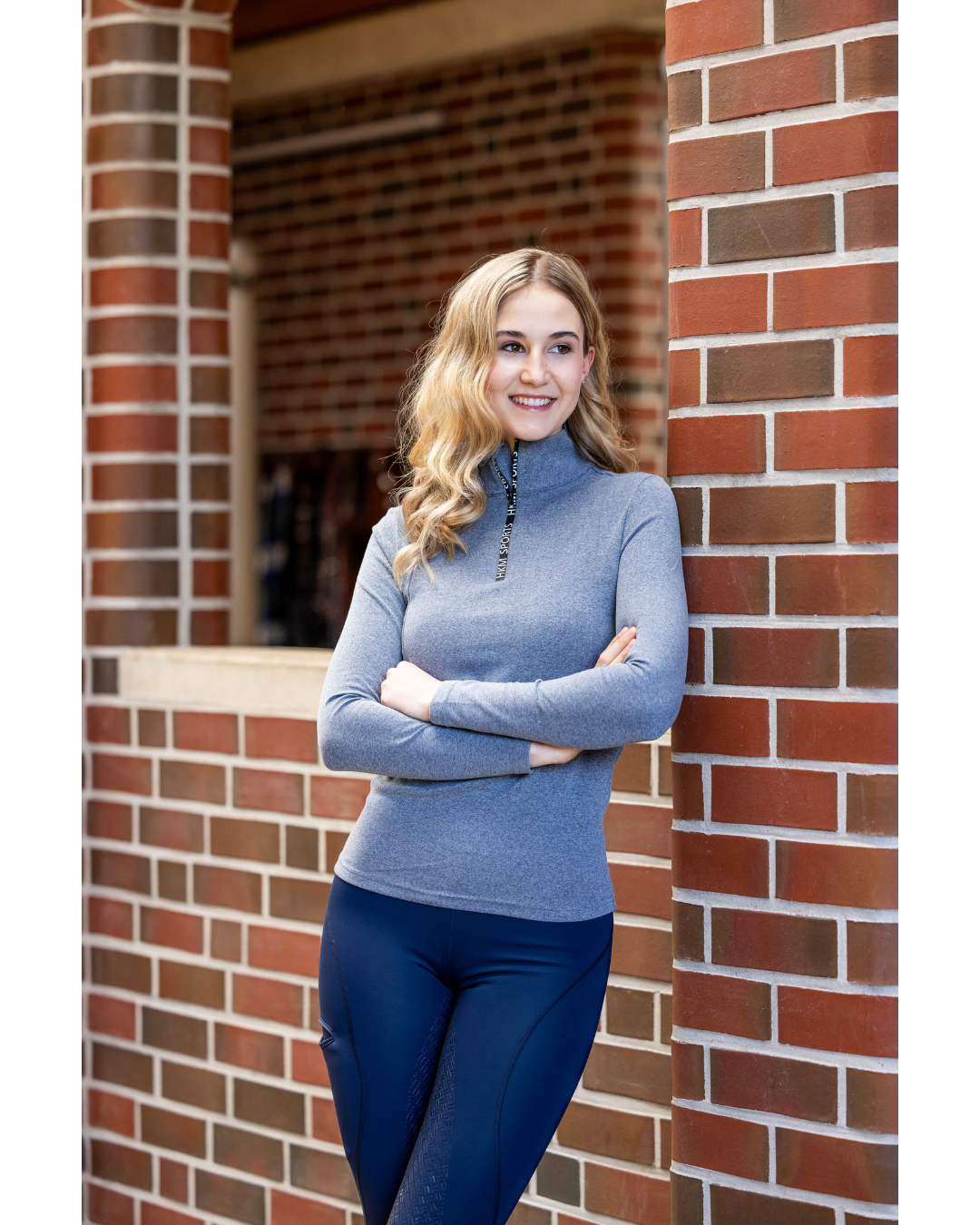 A woman with blonde hair stands leaning against a red brick wall, wearing a heathered blue mock neck  1/4 zip long-sleeve top and dark blue tights equestrian fashion outfitters