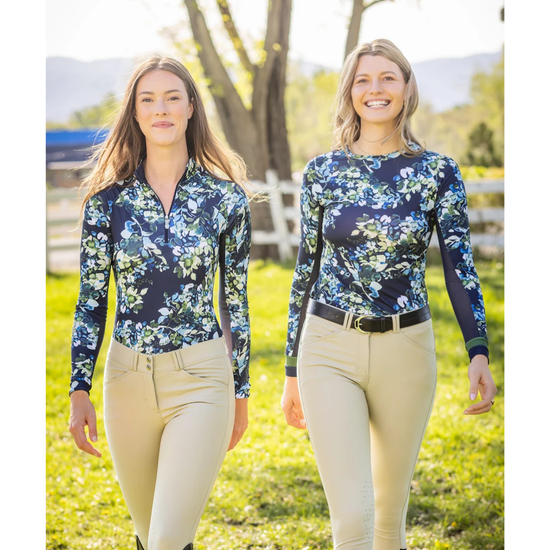 Two women walking outdoors in a grassy field wearing matching long-sleeve navy floral quarter-zip shirts and beige riding pants equestran fashion outfitters
