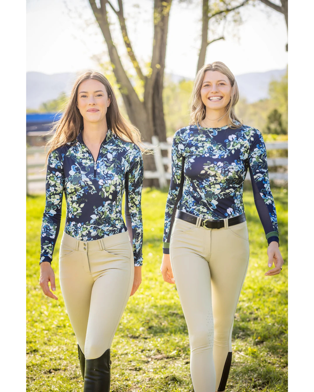 Two women walking outdoors in a grassy field wearing matching long-sleeve navy floral quarter-zip shirts and beige riding pants equestran fashion outfitters