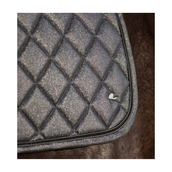 Close-up of a black quilted saddle pad with a diamond pattern and a small white logo, resting on brown fur. equestrian fashion outfitters