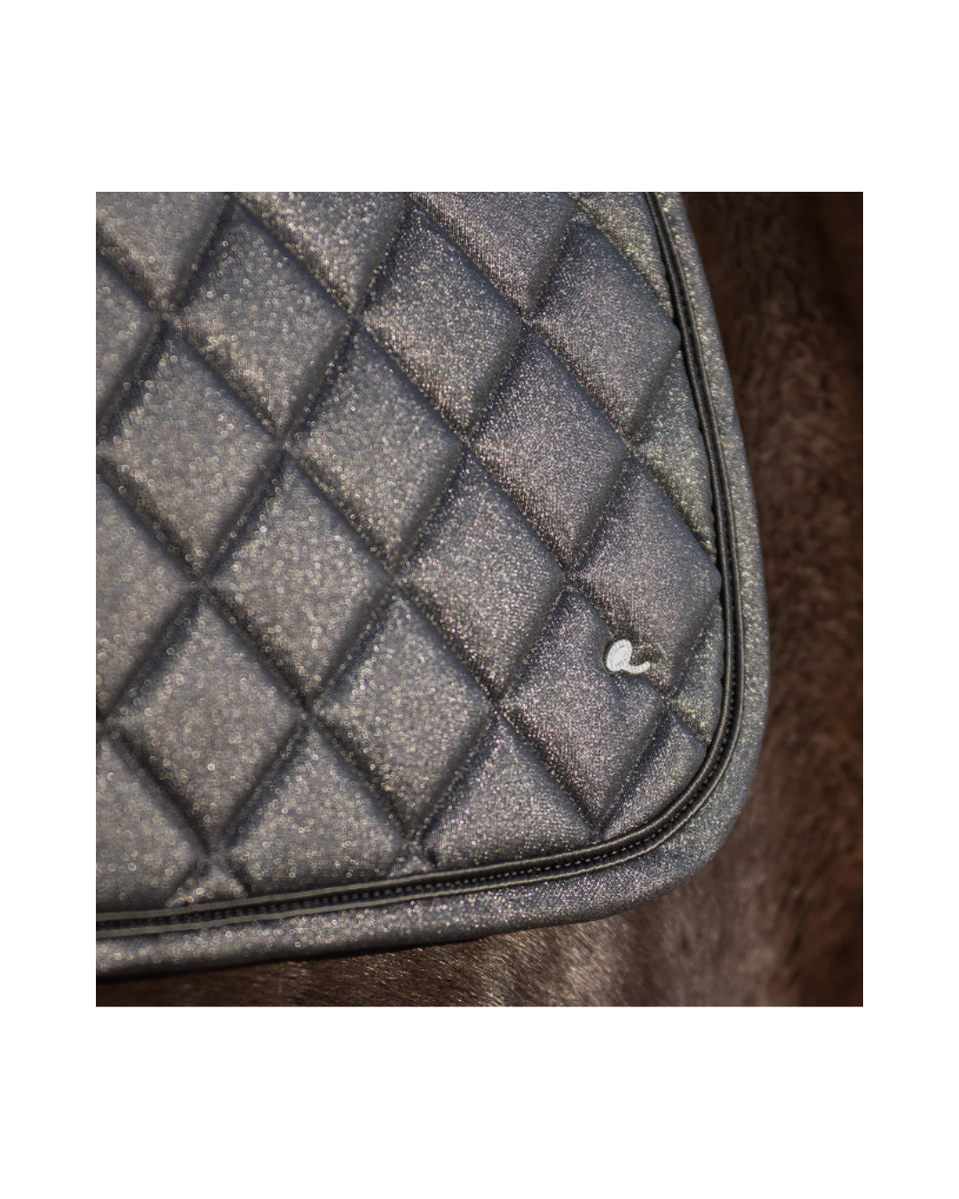 Close-up of a black quilted saddle pad with a diamond pattern and a small white logo, resting on brown fur. equestrian fashion outfitters