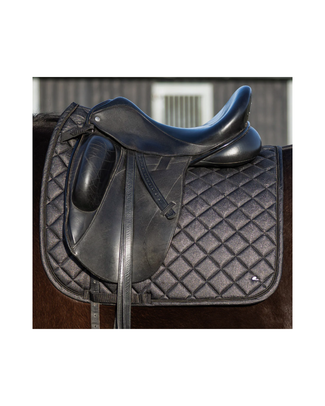 A black leather horse saddle resting on a black diamond-quilted saddle pad, shown against a blurred barn background. equestrian fashion outfitters
