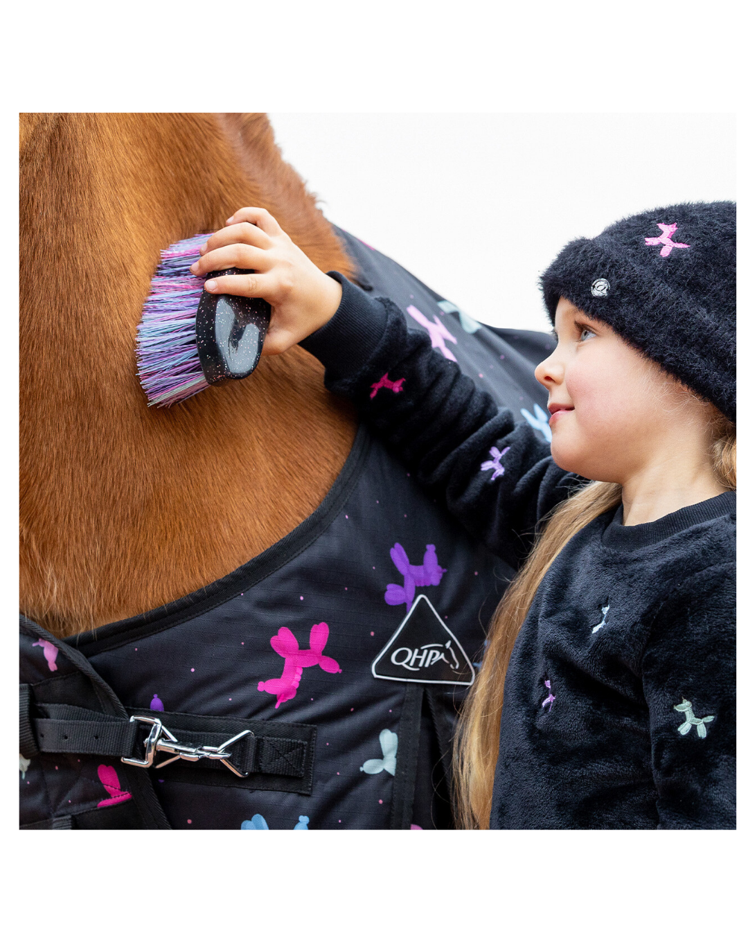 A child wearing a black knit beanie and matching black sweater with colorful motifs gently brushes with a black brush with multi coloured bristles  a brown horse's neck. The horse is wearing a black horse blanket decorated with pink, purple, and blue dog-bone patterns, and a white Q4H! logo is visible on the front.equestrian fashion outfitters