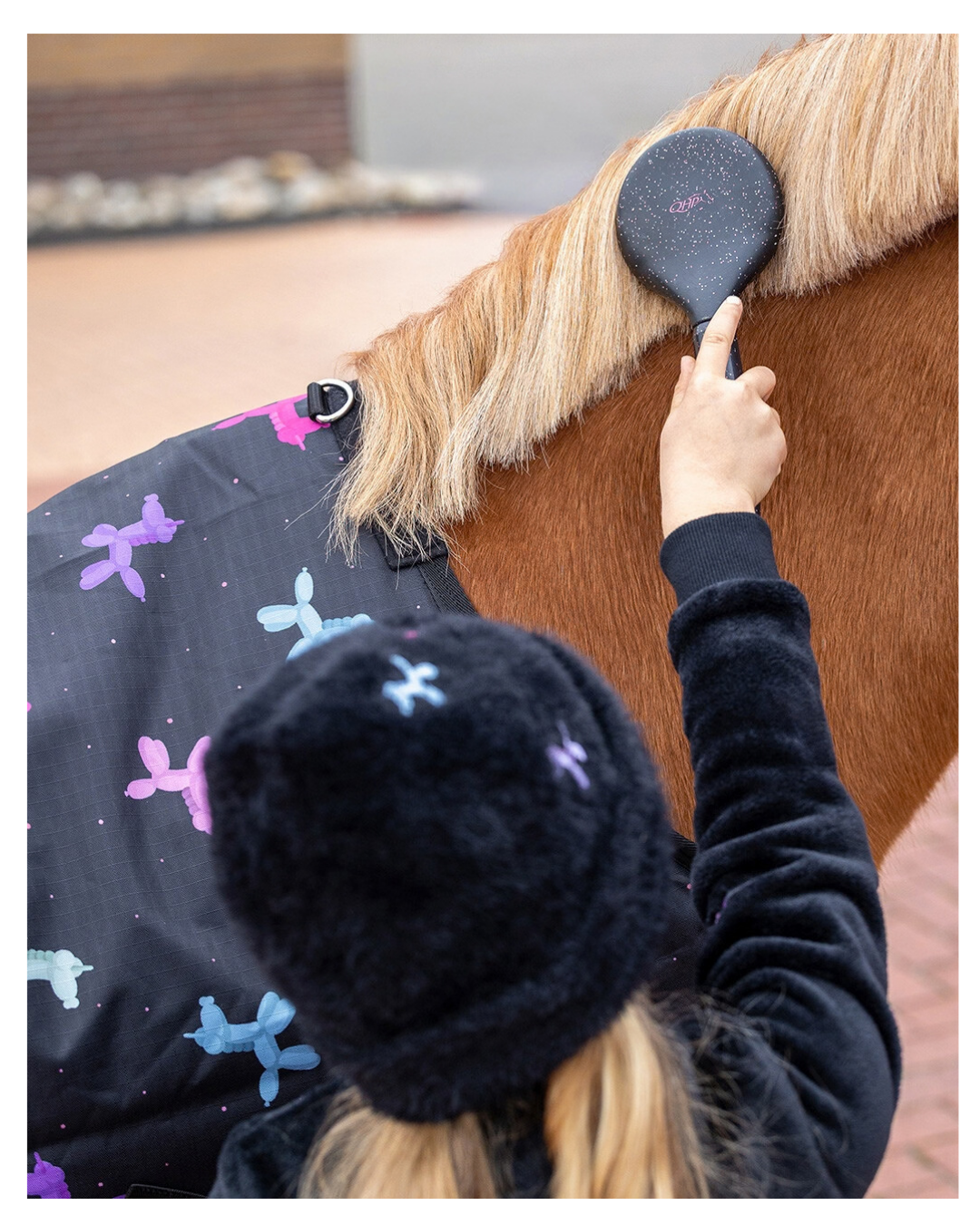 A person wearing a black fuzzy beanie and black fleece jacket brushes a brown horse's mane with a black glitter horse brush. equestrian fashion outfitters